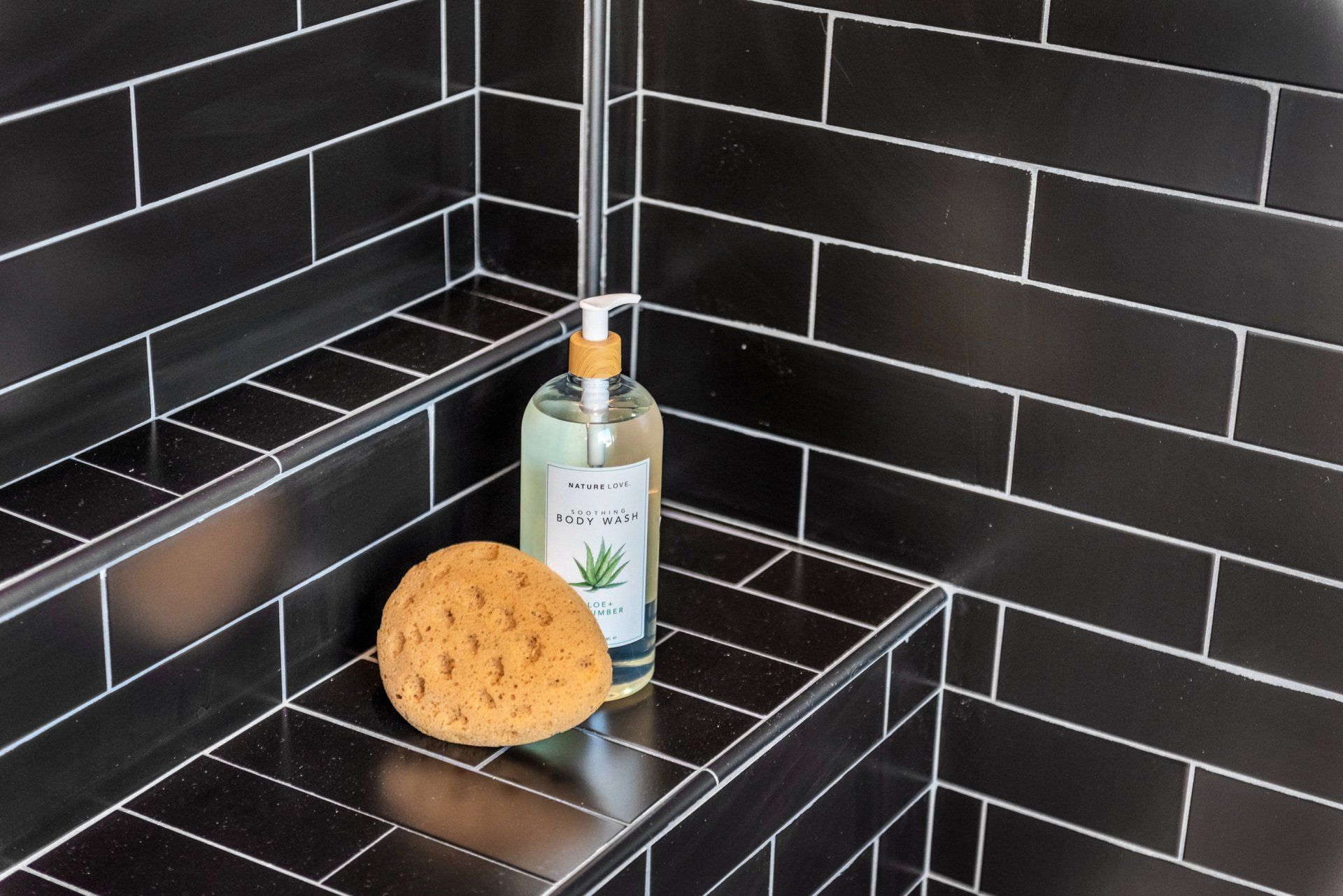 A bottle of soap and a sponge on a black tiled shelf in a shower.