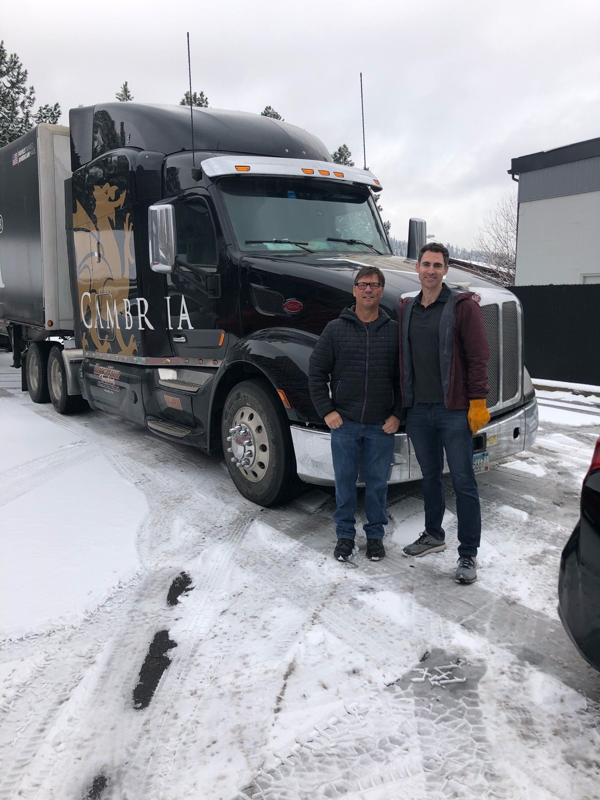 Two men stand by a black semi-truck in a snowy outdoor setting.