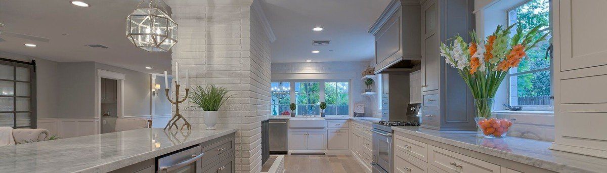 A white kitchen with a lot of cabinets, windows, a brick wall, white countertops, and stainless steel appliances.