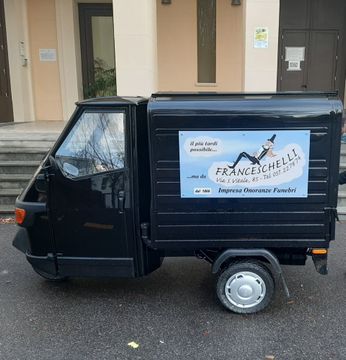 Un piccolo Piaggio Ape nero parcheggiato sul marciapiede, con un cartello pubblicitario 