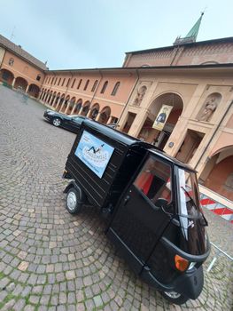 Un piccolo Piaggio Ape nero parcheggiato sul marciapiede, con un cartello pubblicitario 