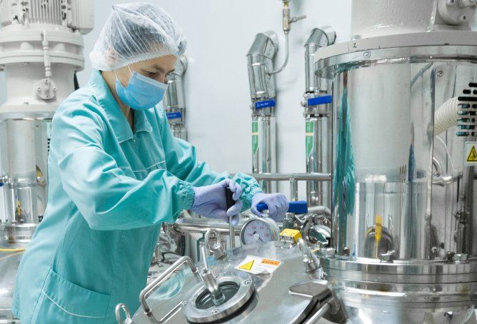 Una persona con bata de laboratorio, mascarilla y redecilla para el pelo ajusta el equipo en una instalación farmacéutica.
