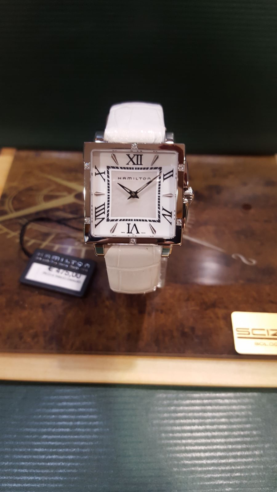 Orologio da polso quadrato con cinturino e quadrante bianchi, esposto su una superficie di legno con un'etichetta con il prezzo.