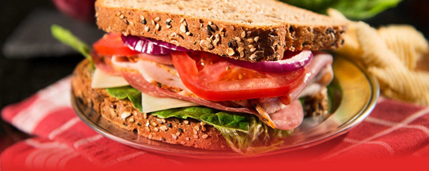 Photo of a sandwich featuring several meats, cheese, lettuce, tomato & red onion on a multigrain bread