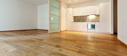An empty room with hardwood floors and a kitchen in the background.