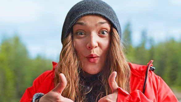 Woman in gray beanie and red jacket smiling with thumbs up outdoors