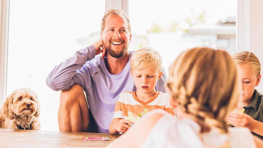 Family gathered at a table, playing a game in a bright living room, with a dog nearby.