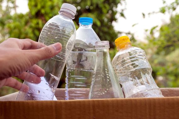 Una mano che getta bottiglie di plastica in un bidone di legno all'aperto, suggerendo il riciclo o la raccolta differenziata dei rifiuti.