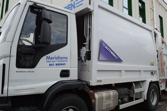 Camion della spazzatura bianco parcheggiato vicino a un edificio, con loghi e scritte aziendali blu sulla cabina e sulla fiancata.