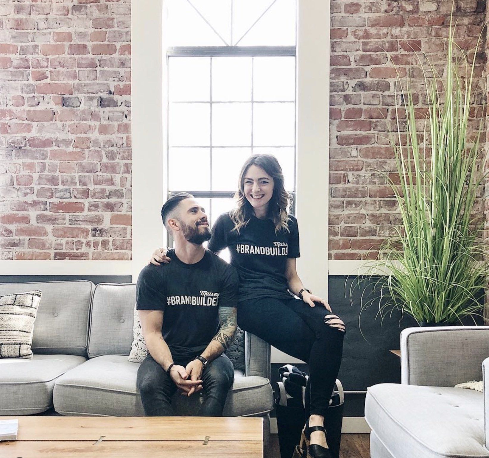 Debbie & Gabriel Mayes sit on a gray couch in their office as they smile about how to grow your business with us at The Mayes Team.
