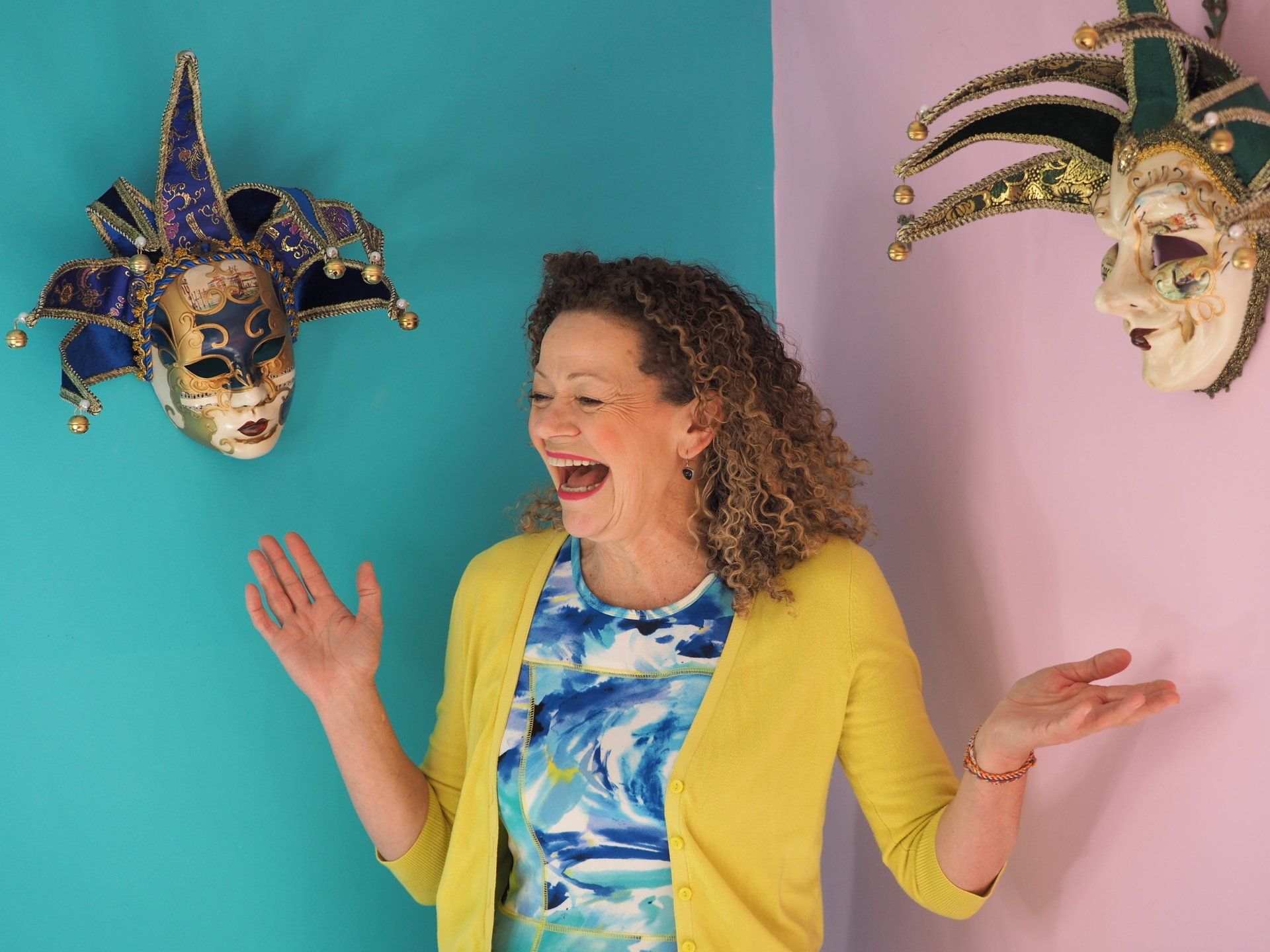 Woman laughing between two colorful masquerade masks on a teal and pink background