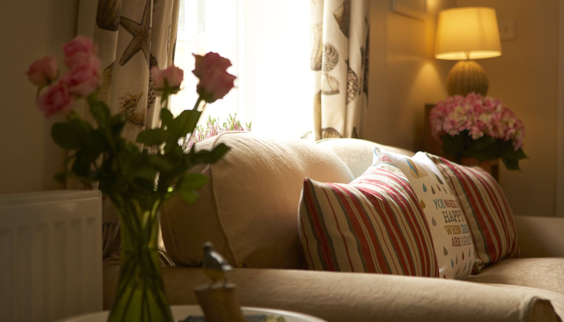 Cozy living room with a sofa, pillows, flowers, and a lamp near a window with sunlight.