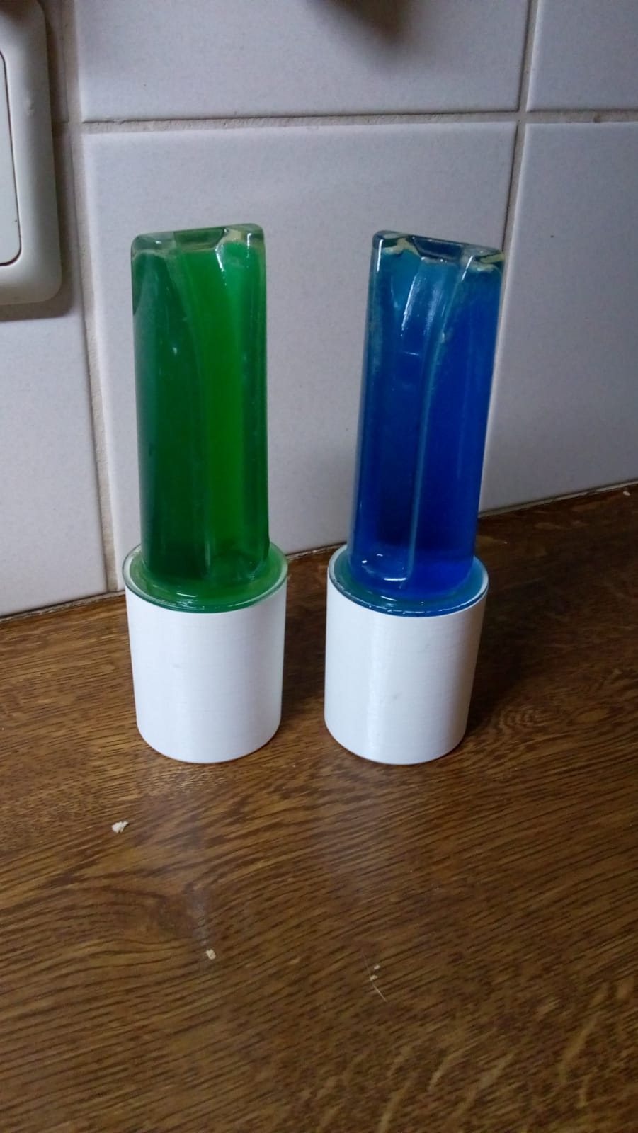 Two colorful toy dispensers, green and blue, standing on a wooden table against tiled wall.