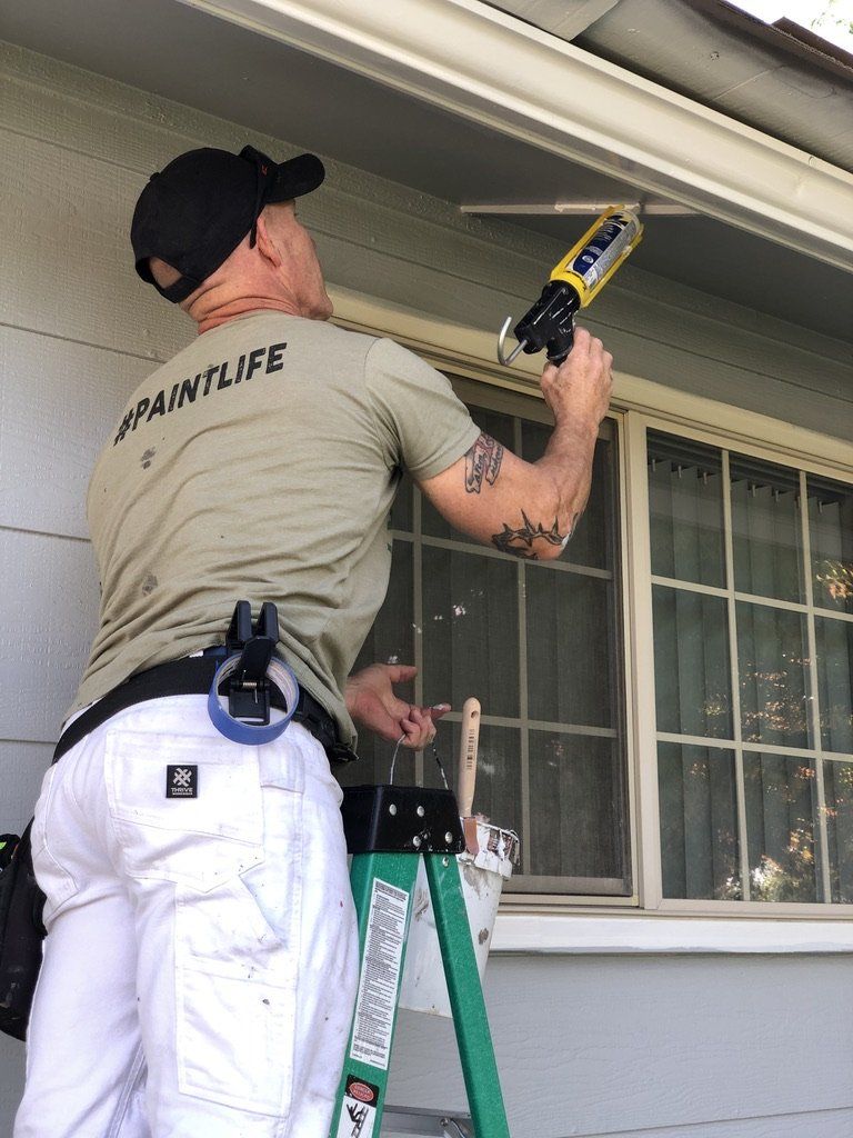 A man is standing on a ladder applying caulk to a window.