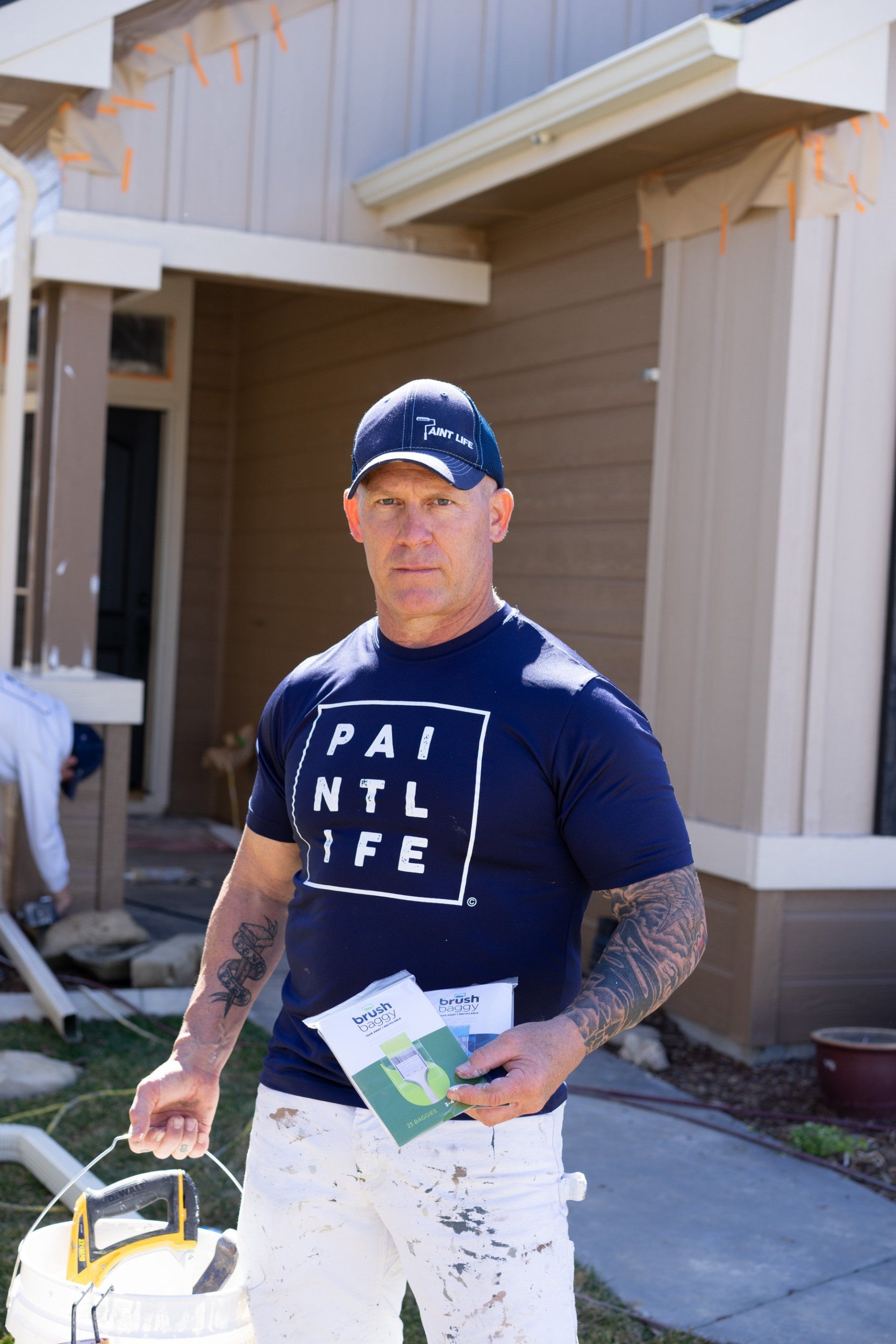 The Idaho Painter wearing a blue shirt that says paint life is standing in front of a house.