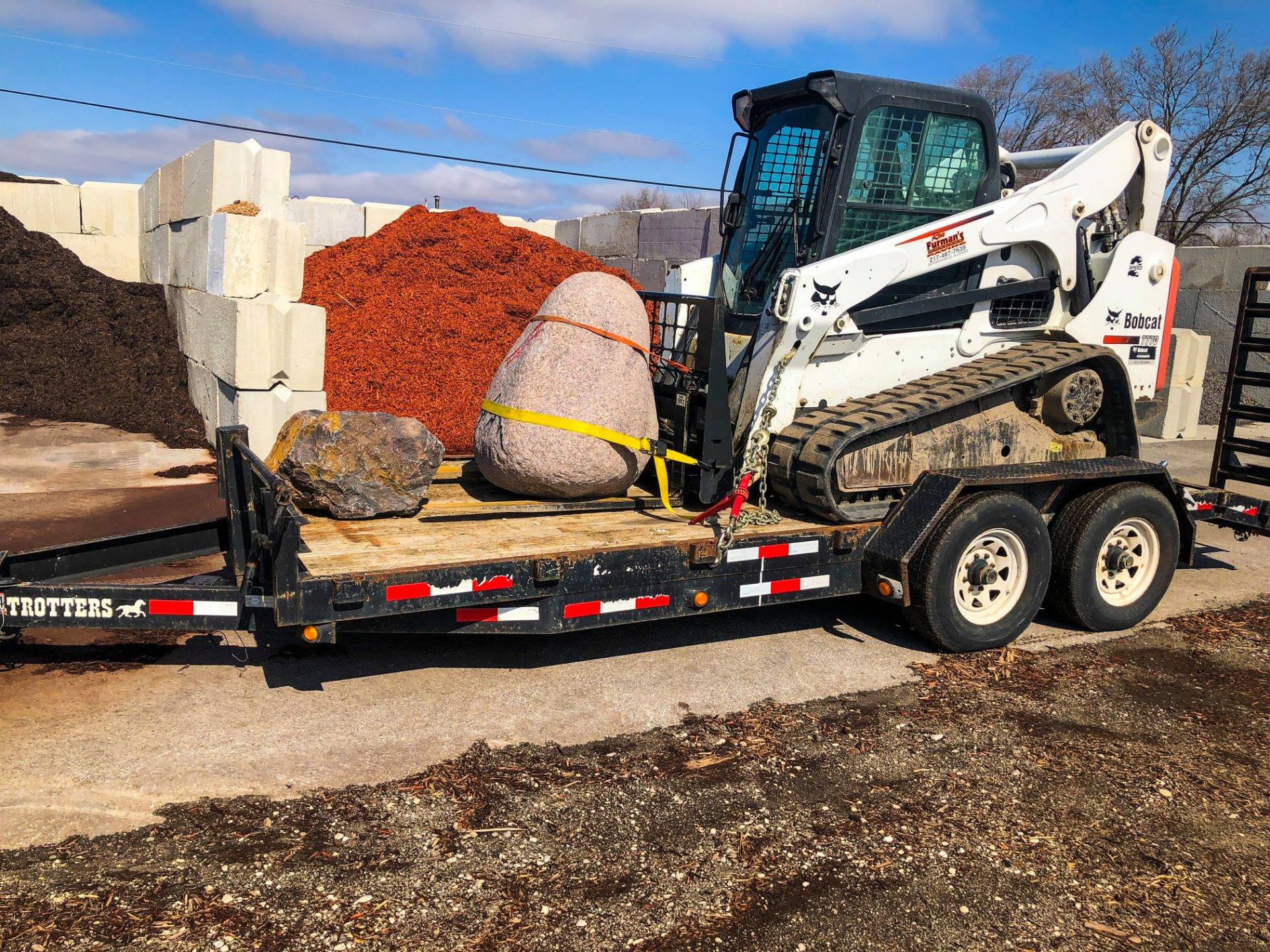 Bobcat skid steer on a trailer hauling rocks and mulch, set against a blue sky.
