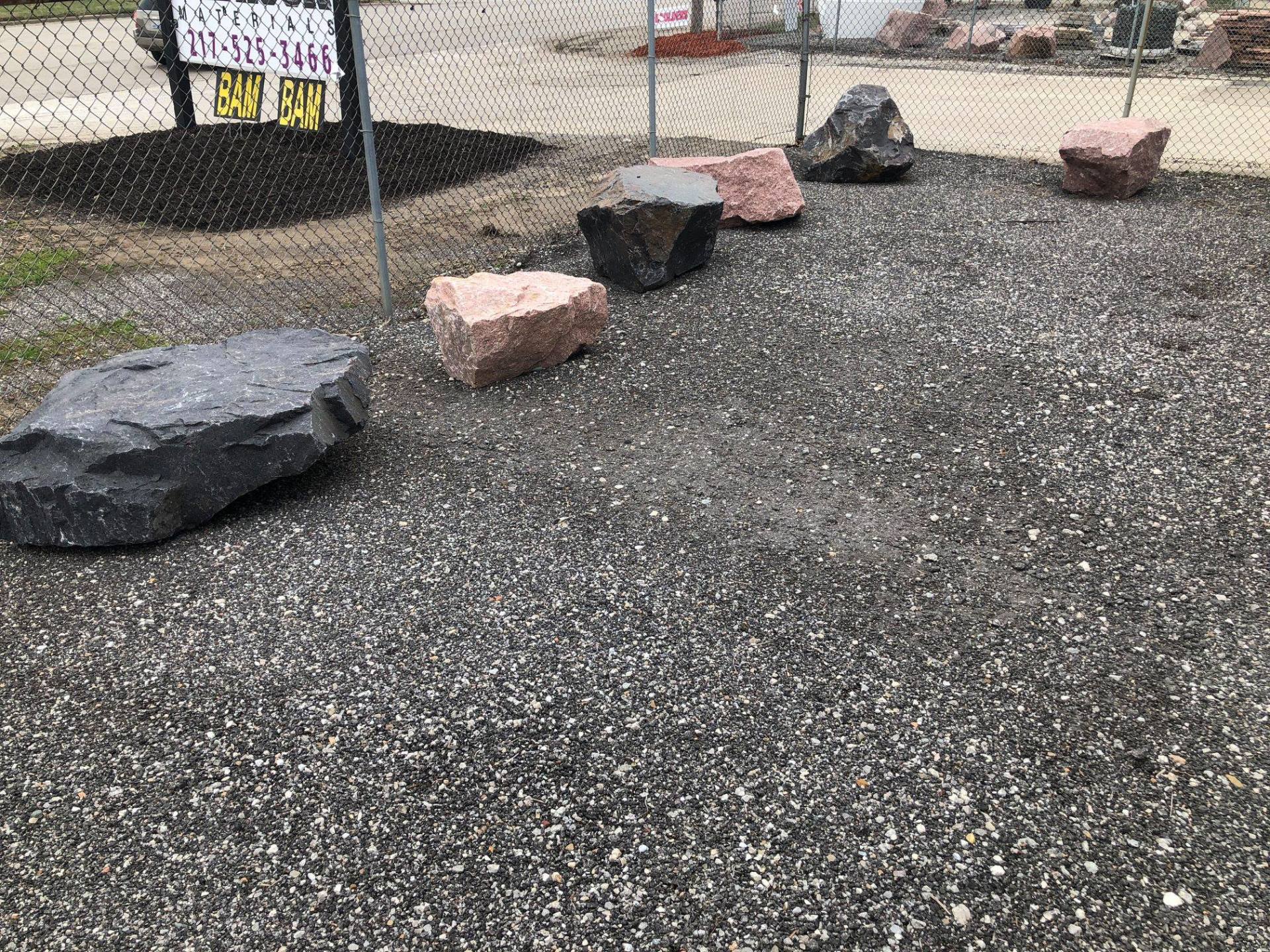 Six large rocks in a gravel-covered area, next to a chain-link fence.
