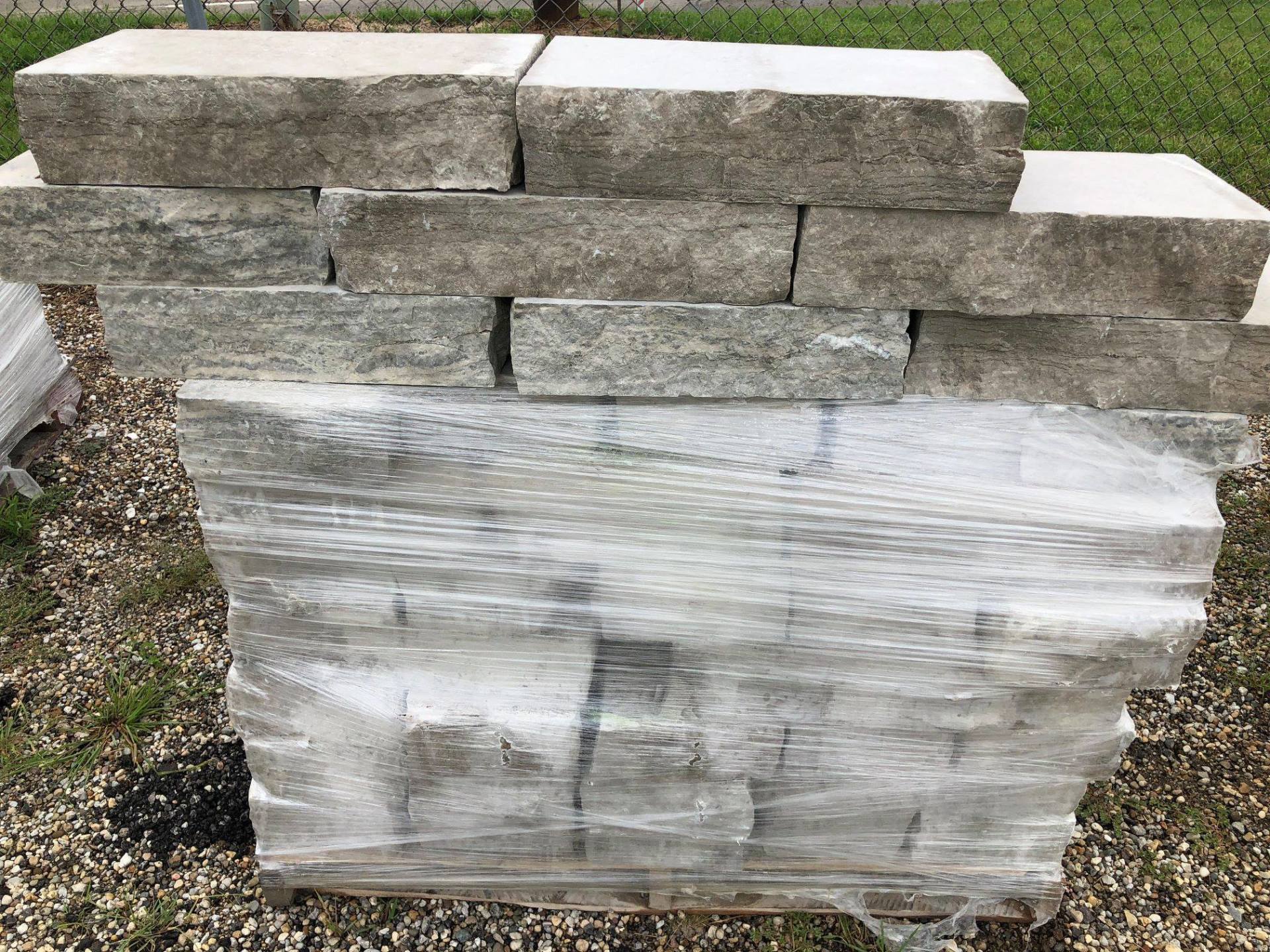 Gray stone bricks stacked, wrapped in plastic, outdoors on gravel.