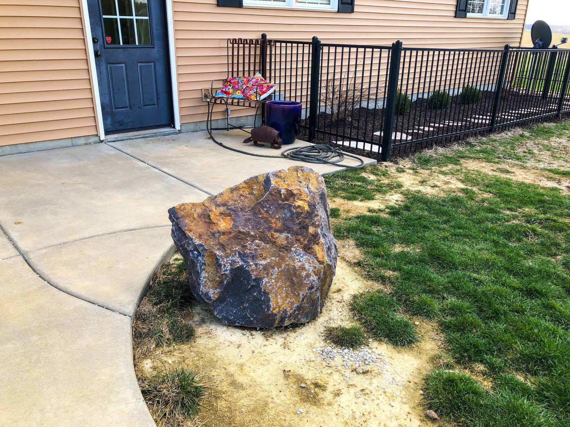 Large rock on a tan sidewalk, near a house with a black door, black fence, and grass.