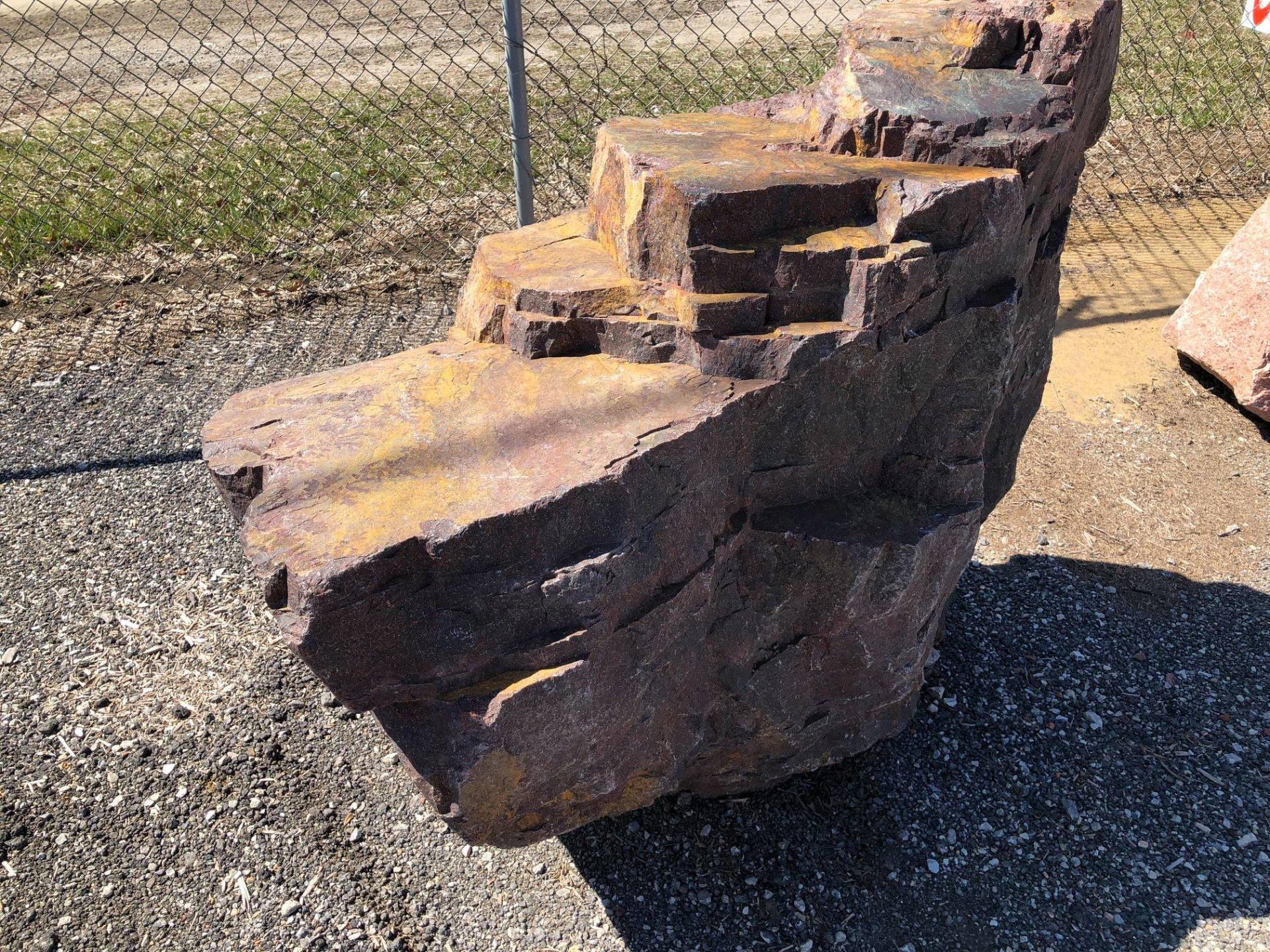 Large, reddish-brown rock with stepped, tiered features, sitting on gravel near a fence.