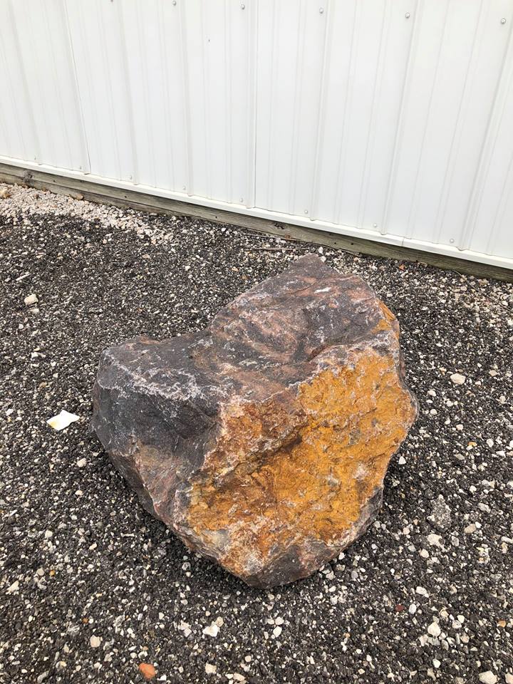 Large rock with brown, black, and orange hues on gravel in front of a white building.