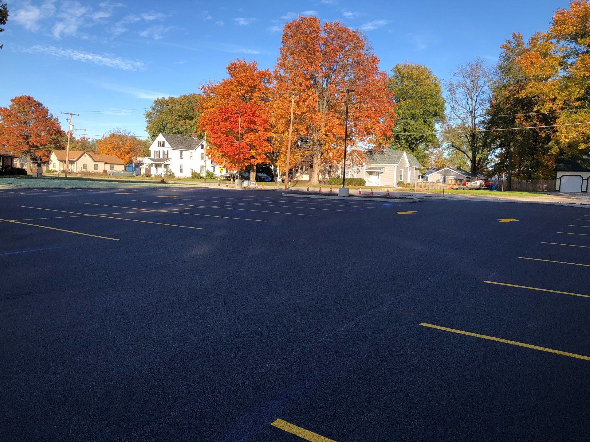 Empty parking lot with trees displaying fall colors; houses in the background under a blue sky.