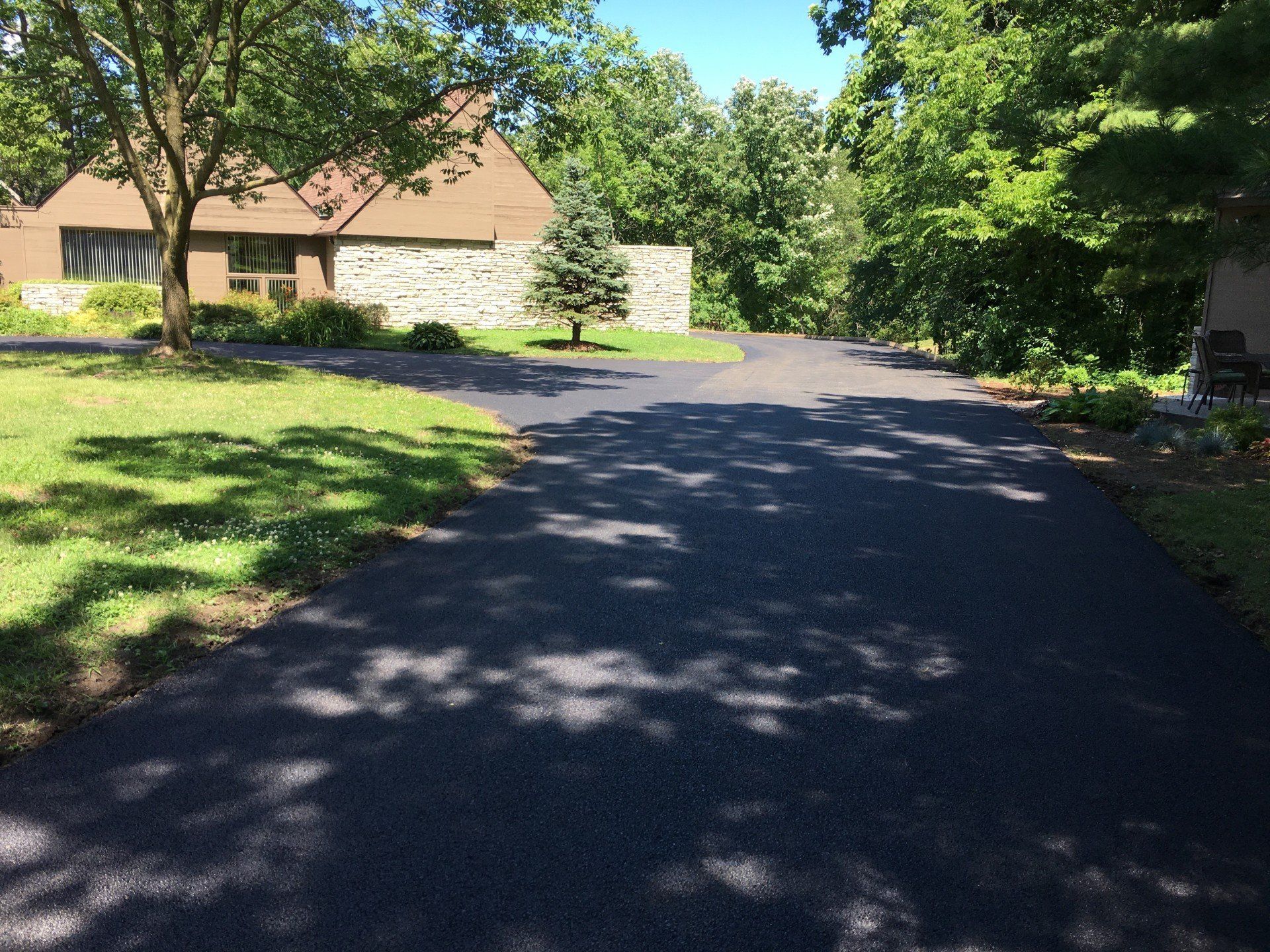 Paved driveway leading to a house with a stone wall; surrounded by trees and green grass on a sunny day.