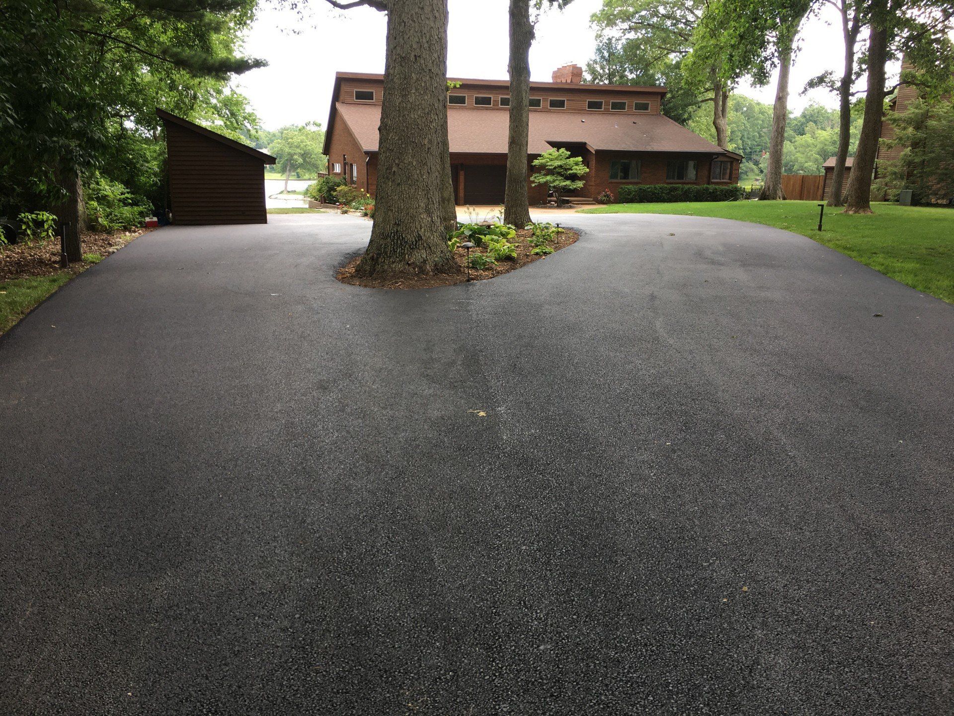 Driveway splits around a tree, leading to a brown house with a green lawn.