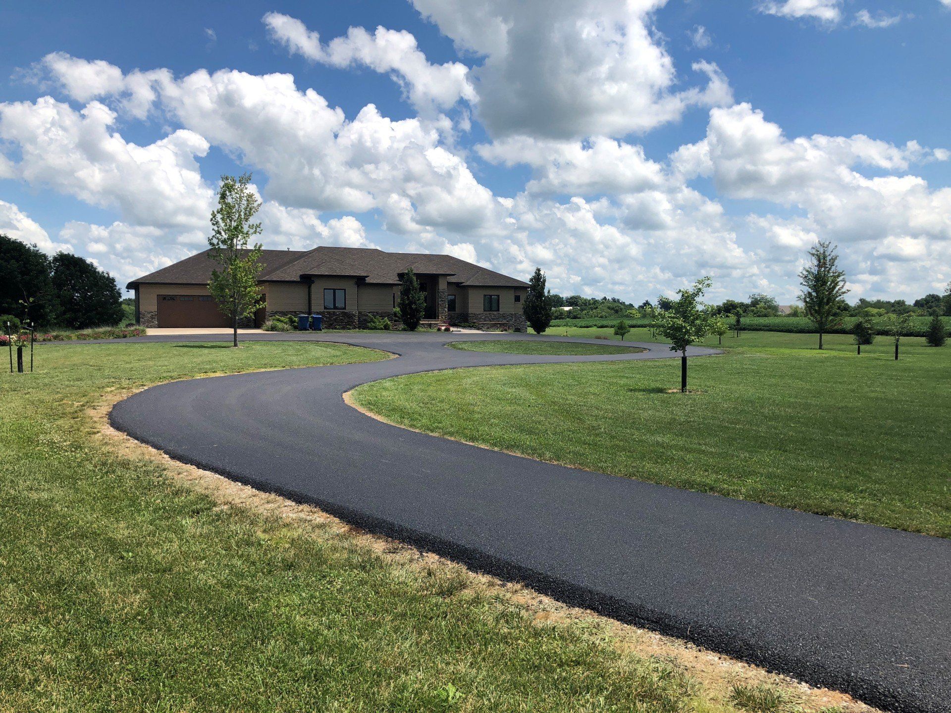 Asphalt driveway curves to a large, tan house on a grassy lawn under a partly cloudy sky.