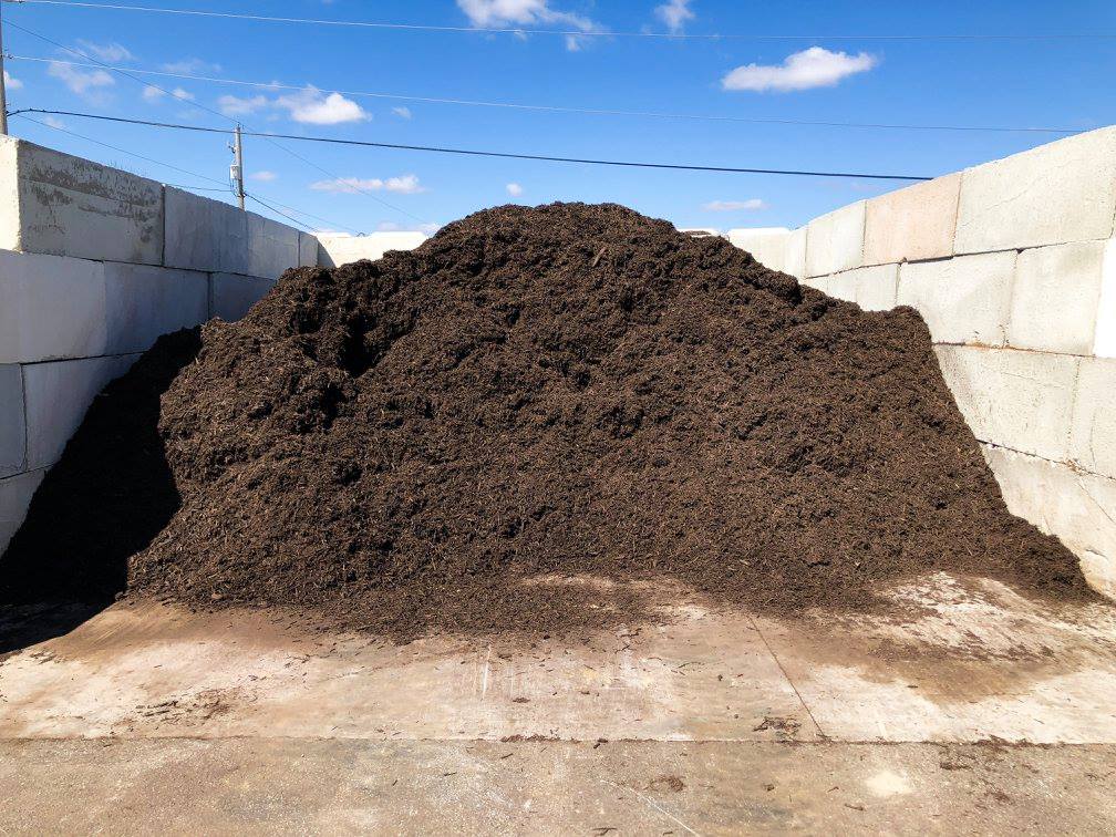 Large pile of dark mulch inside a concrete walled area on a sunny day.