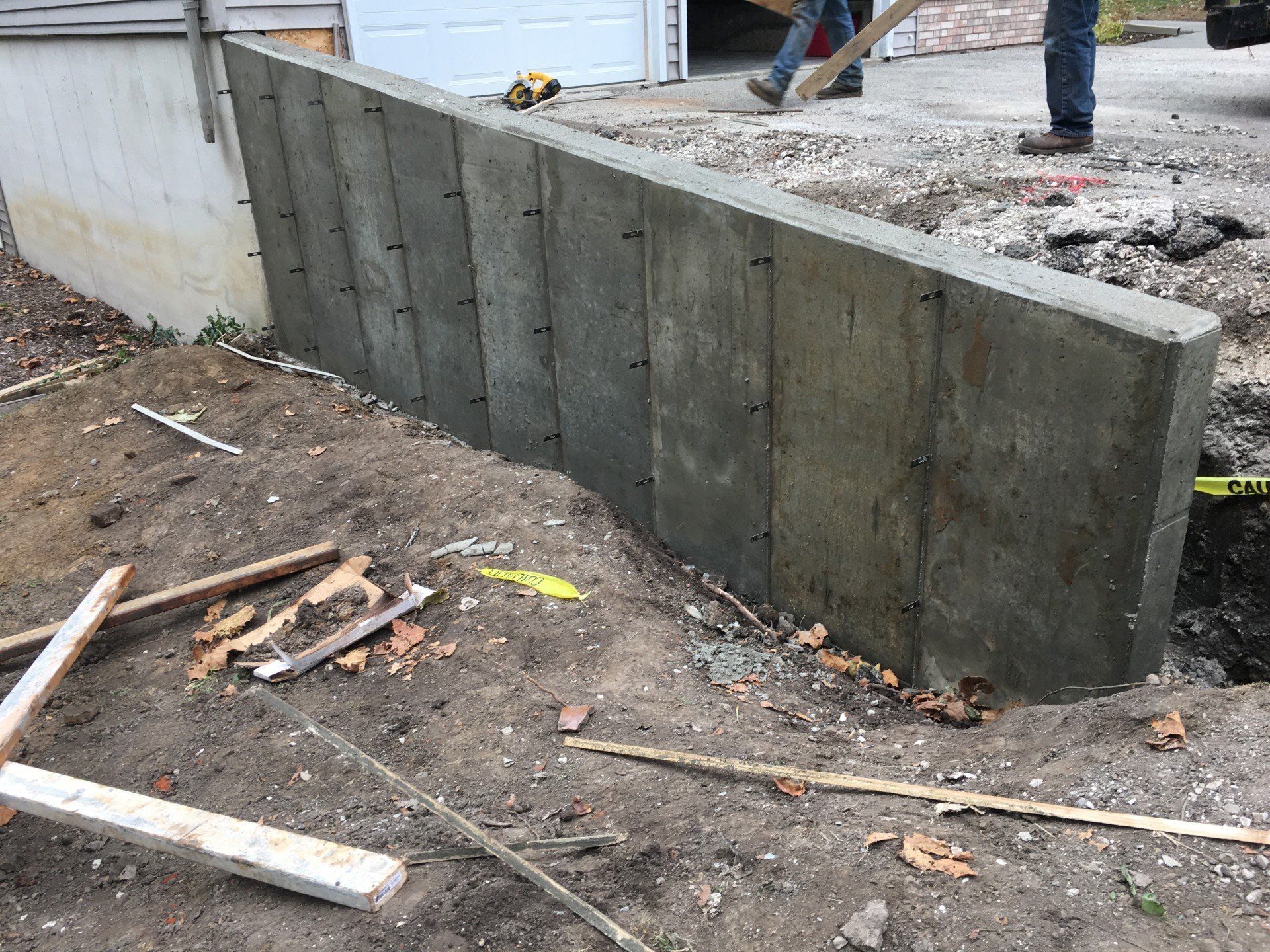 Concrete retaining wall being built beside a garage. Workers, tools, and dirt visible.