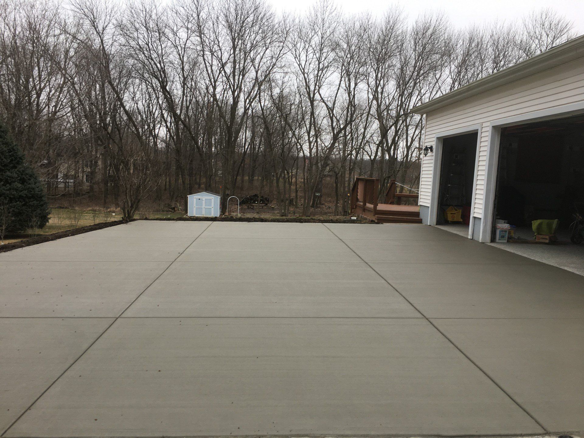 Concrete driveway in front of a garage, a small white storage shed in the background, trees, overcast sky.
