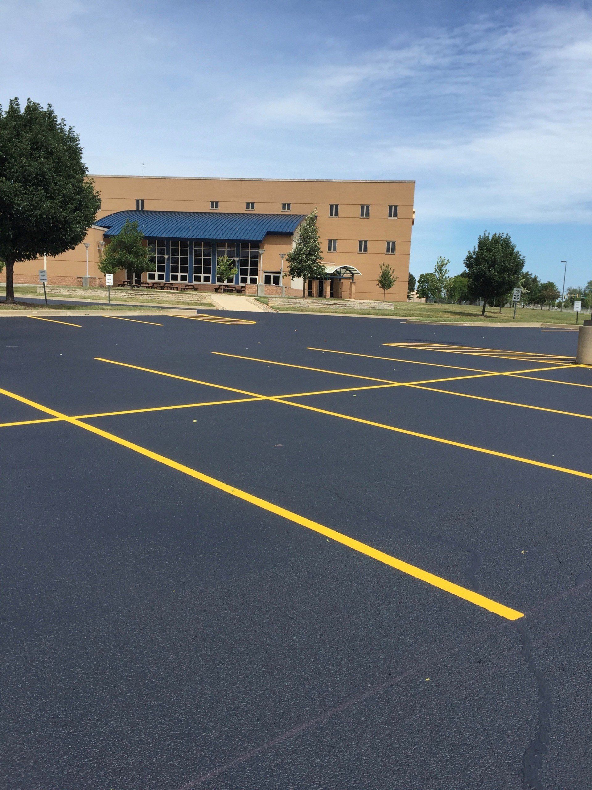 Paved parking lot in front of a tan brick building with a blue roof, under a partly cloudy sky.
