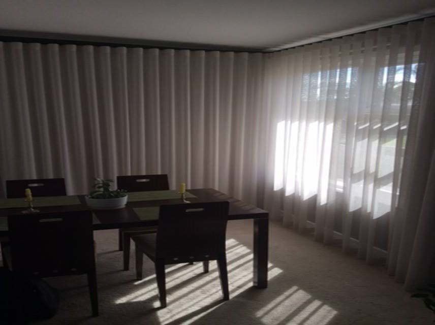 Dining room with a table, chairs, and light-filtering curtains. Sunlight streams through a window.