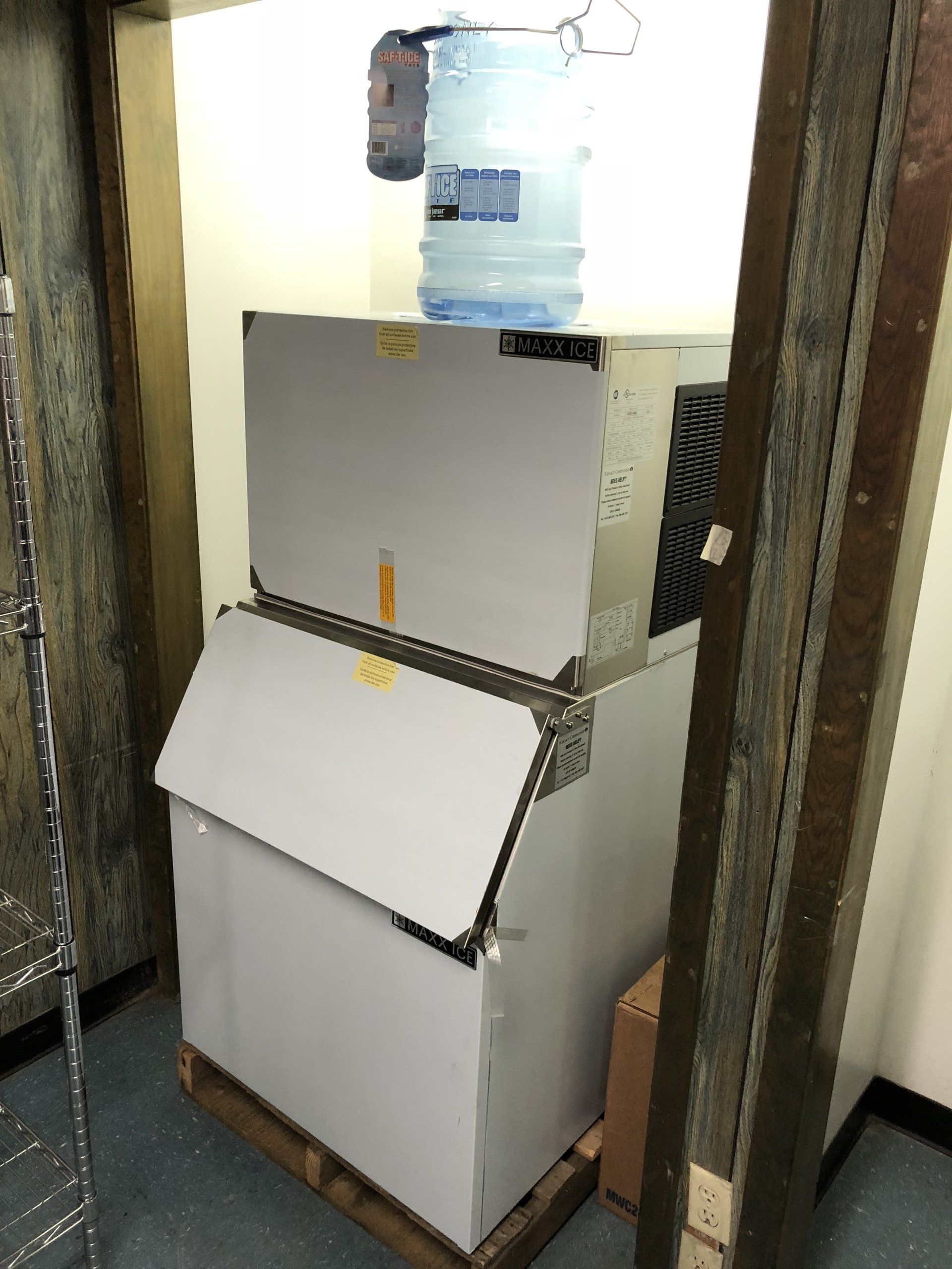 An ice machine is sitting on a wooden pallet in a room next to a water cooler.