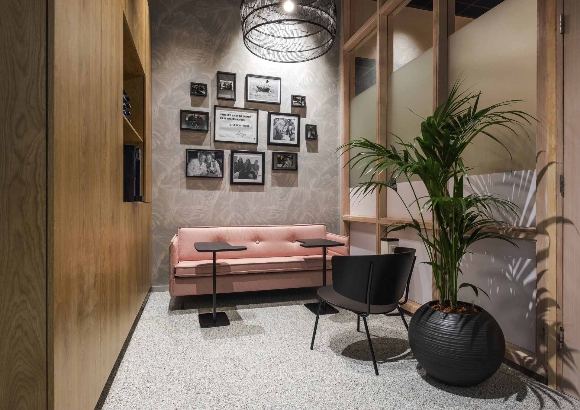 Een kleine zithoek met een roze bank, een zwarte fauteuil, twee bijzettafels, een fotowand en een grote potplant op de vloer in het callcenter van Gemeentehuis van Rijswijk, interieurontwerp door KuóStudio