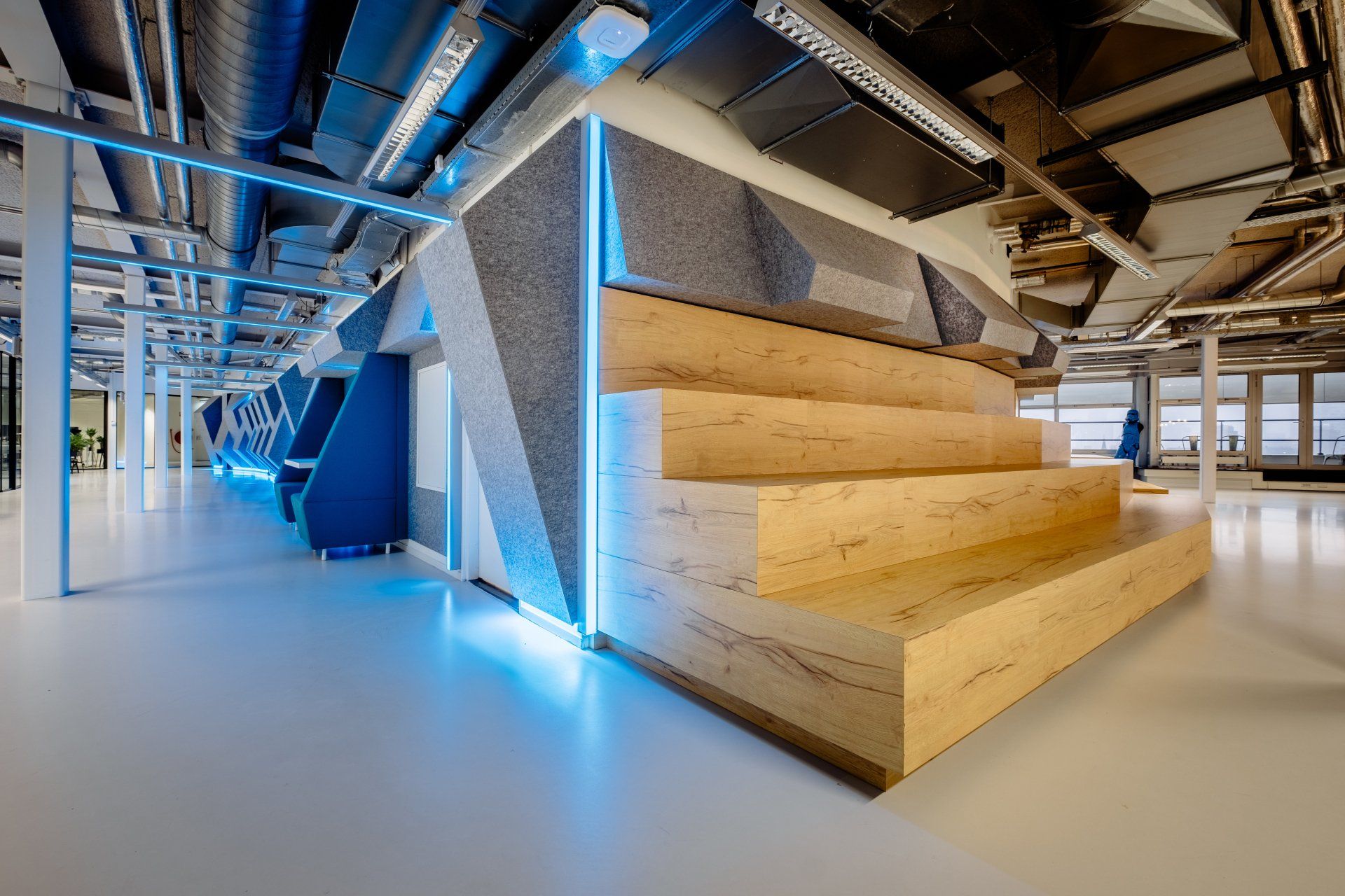 Een modern vitaal kantoorinterieur met een houten zithoek in verschillende niveaus, geaccentueerd met blauwe ledverlichting en geometrische panelen in het nieuwe kantoor van Shypple in Rotterdam, interieurontwerp voor KuóStudio