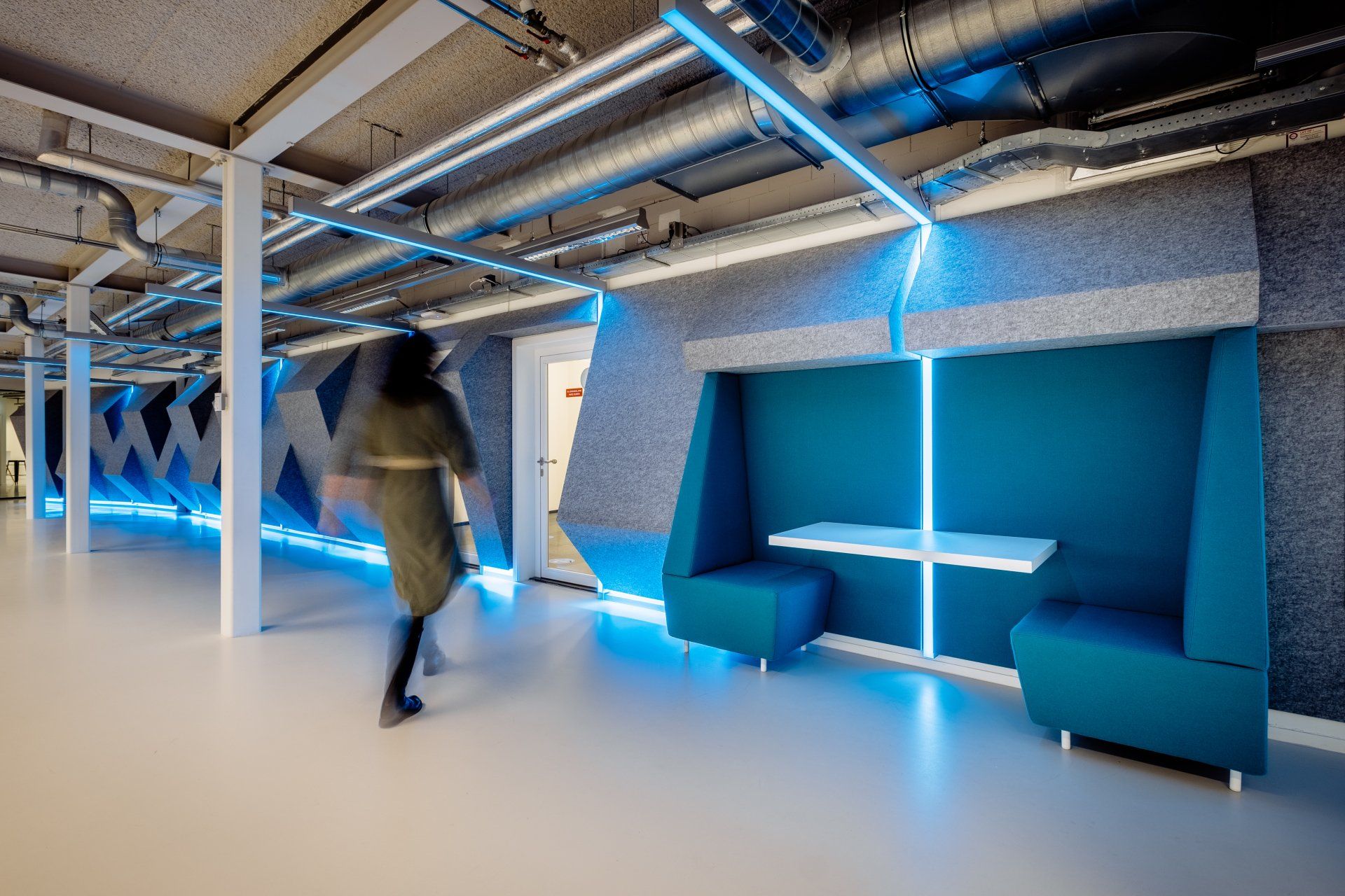 Iemand loopt langs een akoestische blauw verlichte, moderne zithoek in een kantoorruimte met een industrieel, open plafond in het nieuwe kantoor van Shypple in Rotterdam, interieurontwerp voor KuóStudio