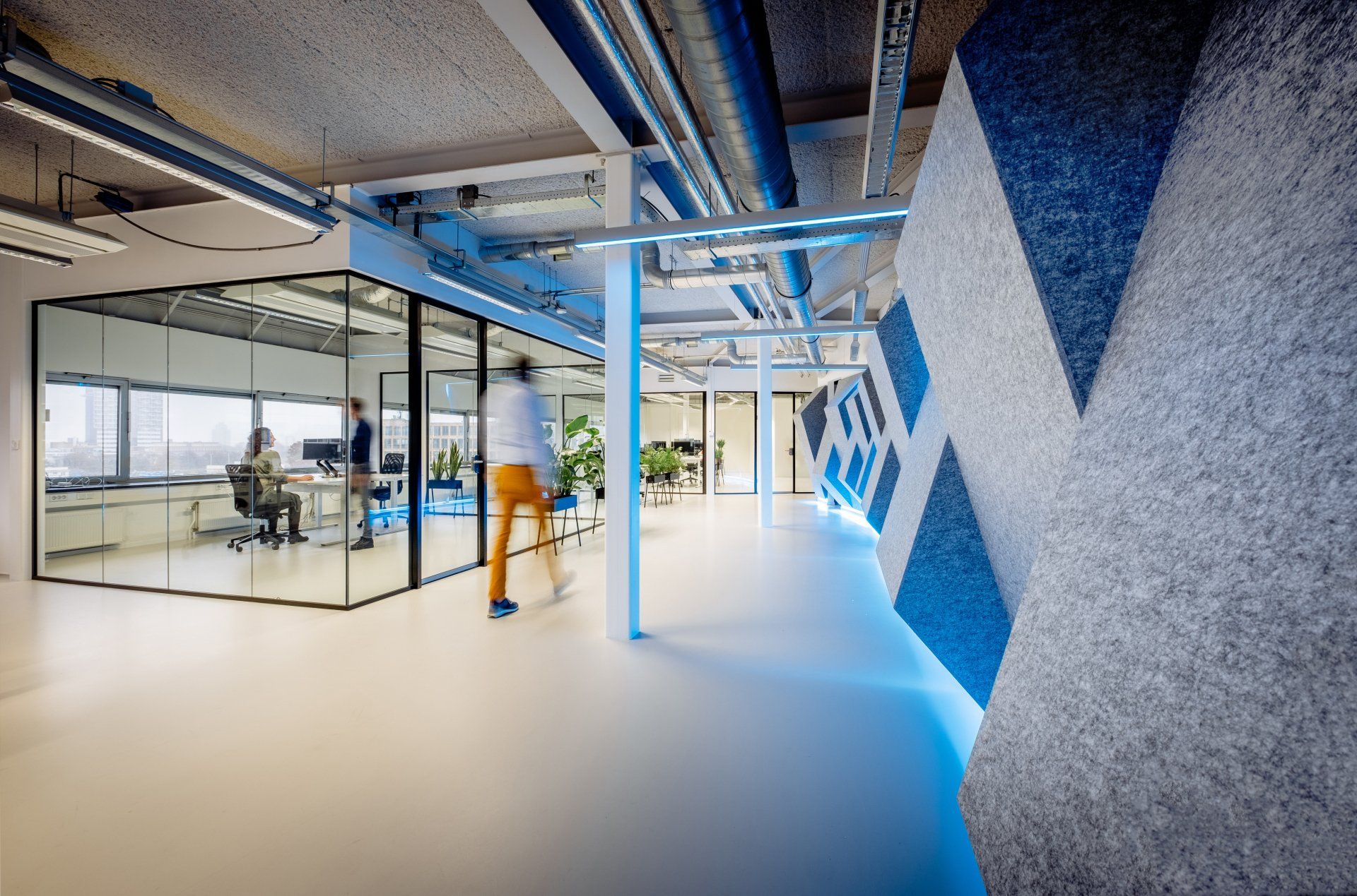 Een modern kantooromgeving met een hoekige grijze muur met 3D textuur, indirecte verlichting, een glazen vergaderruimte en een persoon die rondloopt in het nieuwe kantoor van Shypple in Rotterdam, interieurontwerp voor KuóStudio