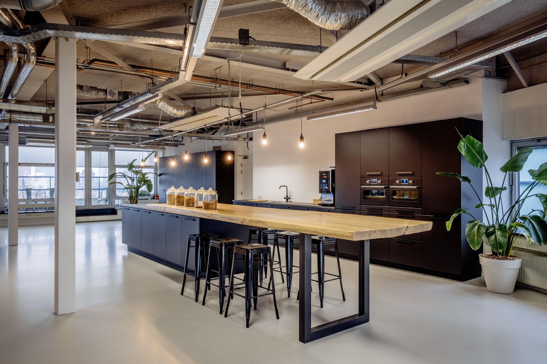 Moderne kantoorkeuken met een grote houten tafel op zwarte metalen poten, omringd door krukken, in het open industriële werkcafé in het nieuwe kantoor van Shypple in Rotterdam, interieurontwerp voor KuóStudio
