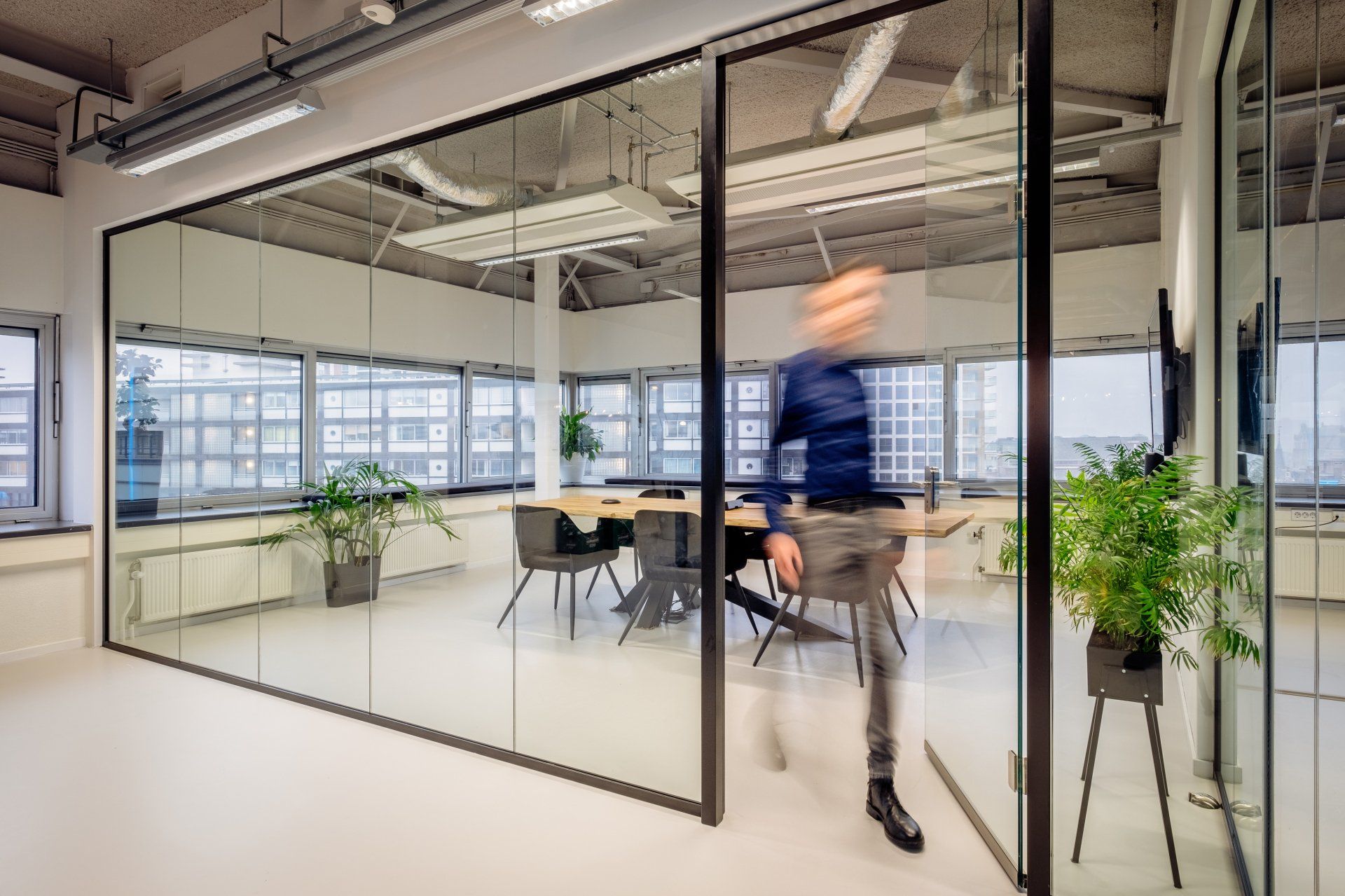 Een persoon loopt door een glazen vergaderruimte met een houten tafel en planten in het nieuwe kantoor van Shypple in Rotterdam, interieurontwerp voor KuóStudio