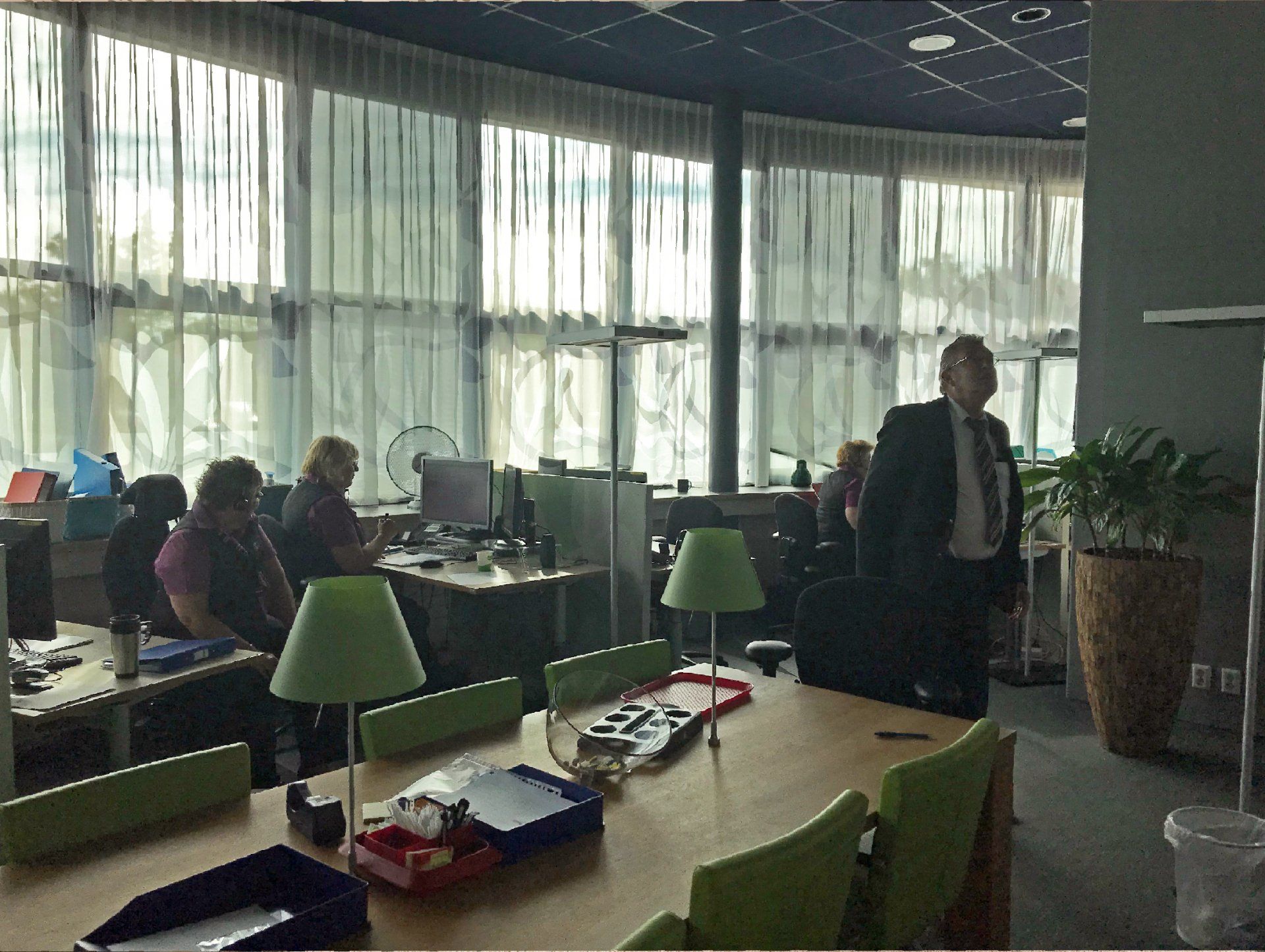 Voor verbouwing: een callcenter met bureaus, groene stoelen, een vergadertafel en een persoon die op de voorgrond staat in klant contact centrum van het Gemeentehuis van Rijswijk