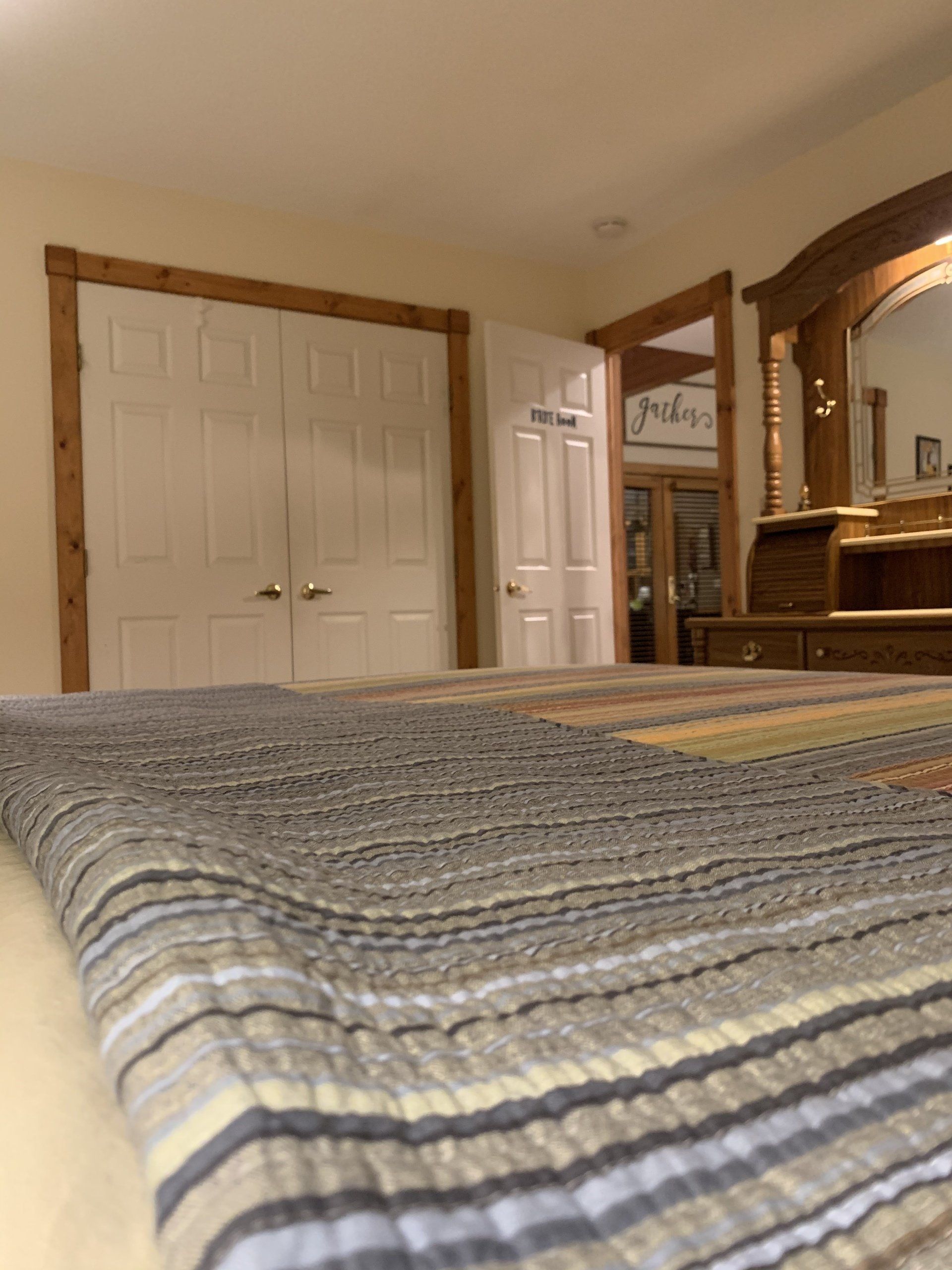 Bedroom with a bed in the foreground, closet and vanity in the background. Quilt is striped.