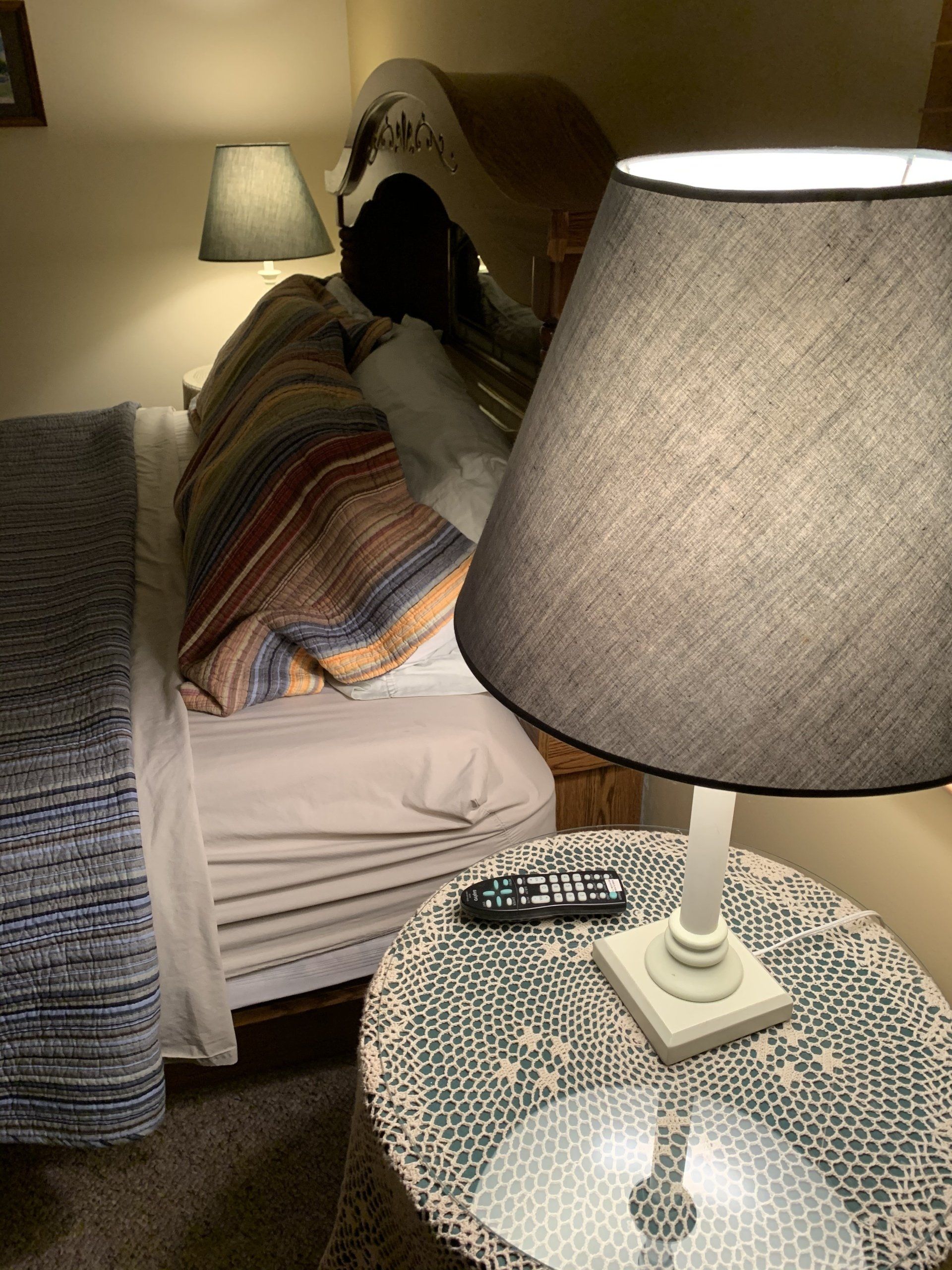 Bedroom scene. Bed with colorful blanket, bedside table with lamp and remote.  Two lamps illuminated.