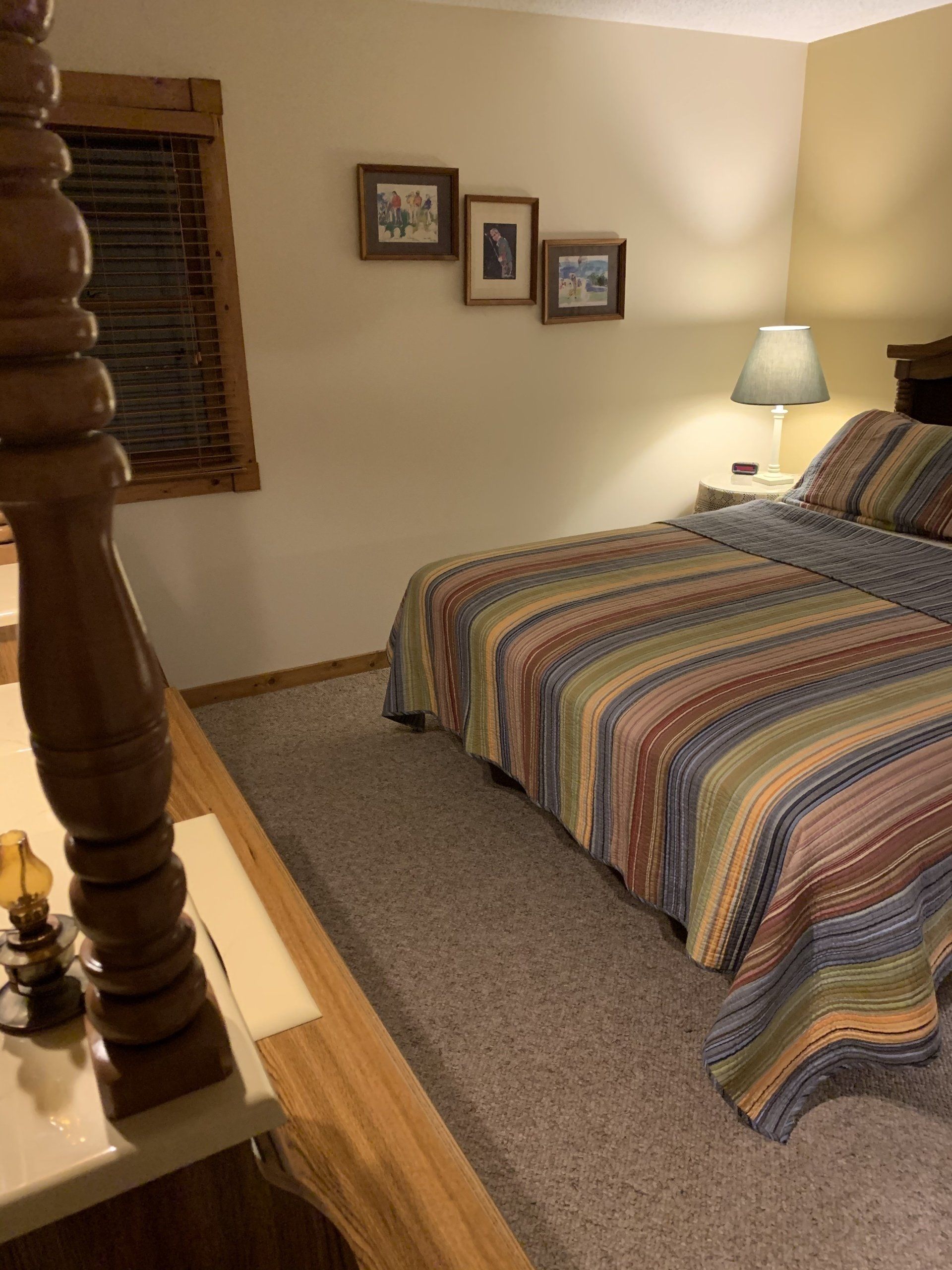 Bedroom with bed, striped quilt, artwork, lamp, and wooden blinds.