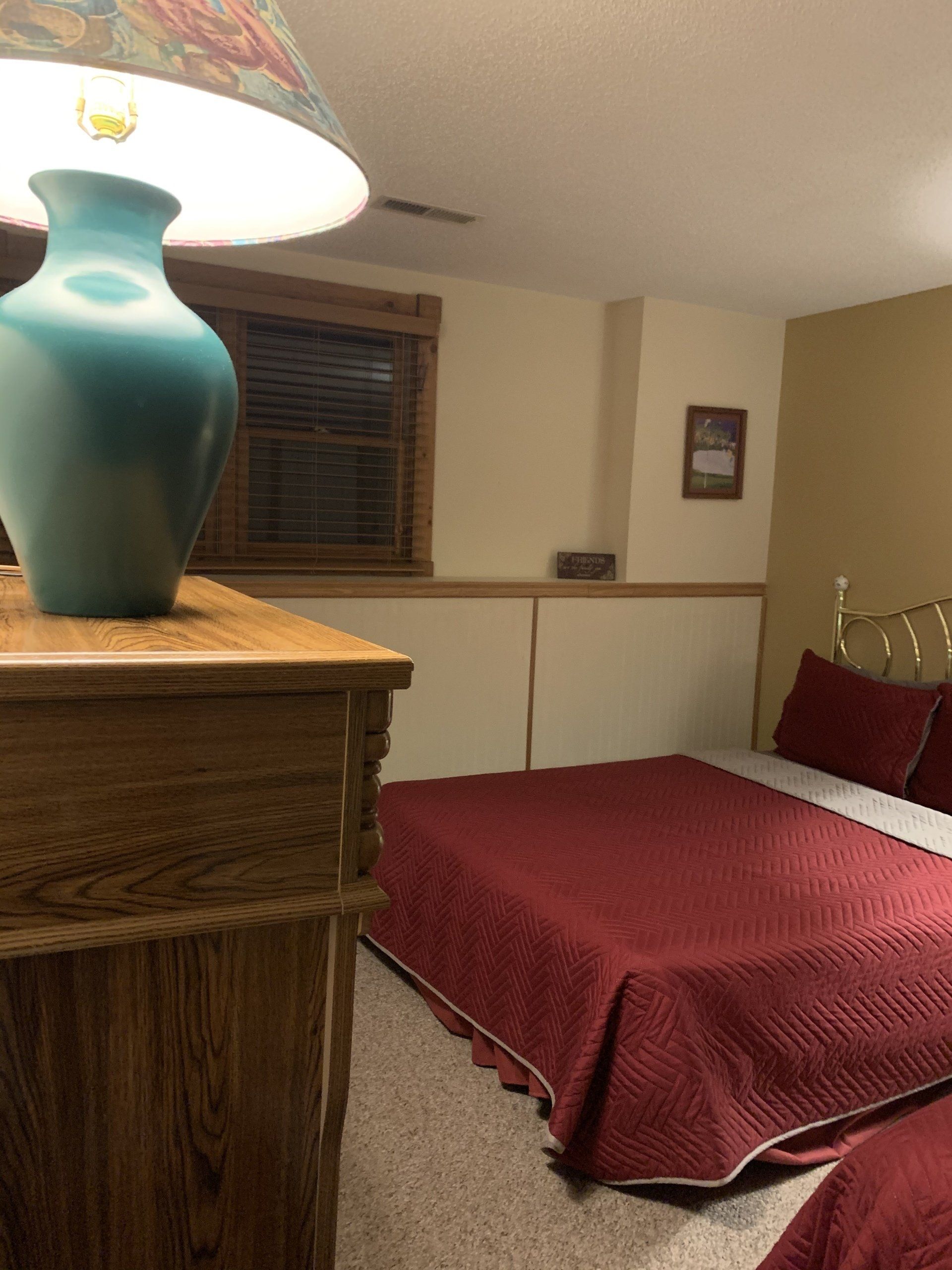 Bedroom with two beds covered in red quilts, a wood dresser with a teal lamp, and a small window.