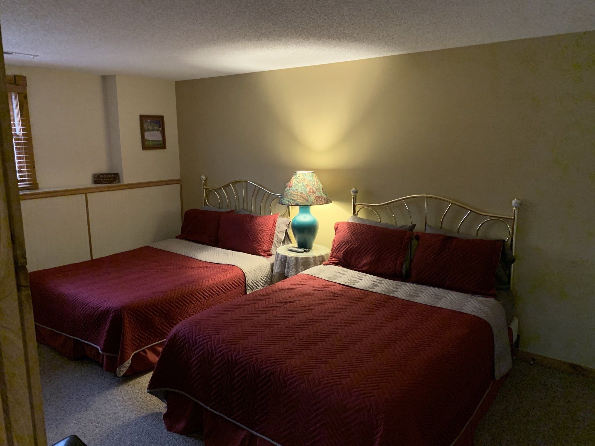 Bedroom with two double beds, red bedding, a small table with a lamp, and tan walls.