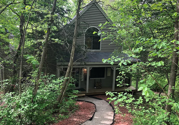 Cabin nestled in trees with a stone path leading to the porch. Gray siding, arched window.