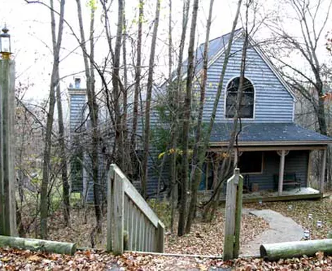 House with wooden siding and porch, partially obscured by bare trees and a wooden staircase.
