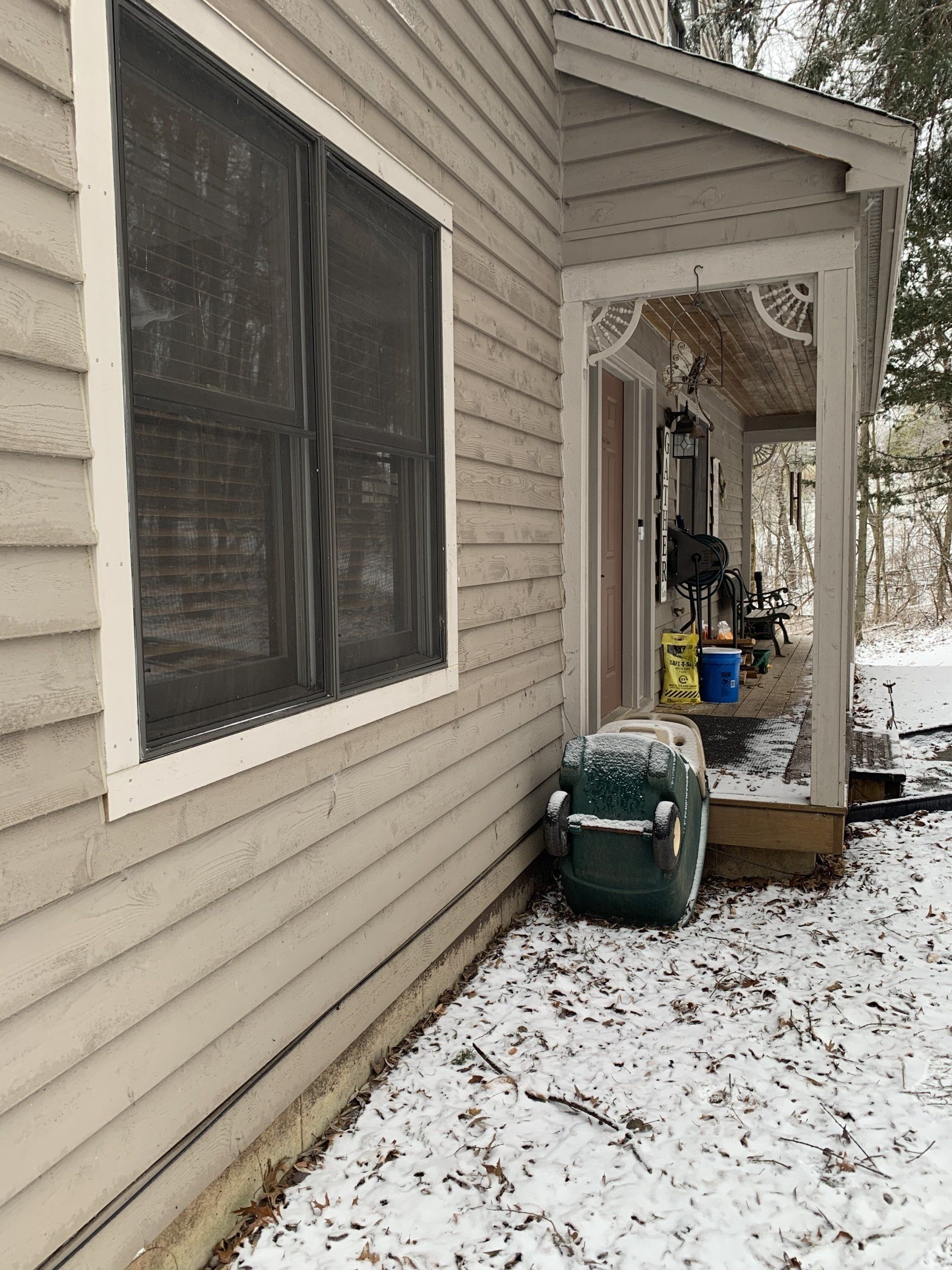 Exterior view of a house with a snow-covered porch, window with a screen, and a green object in the foreground.